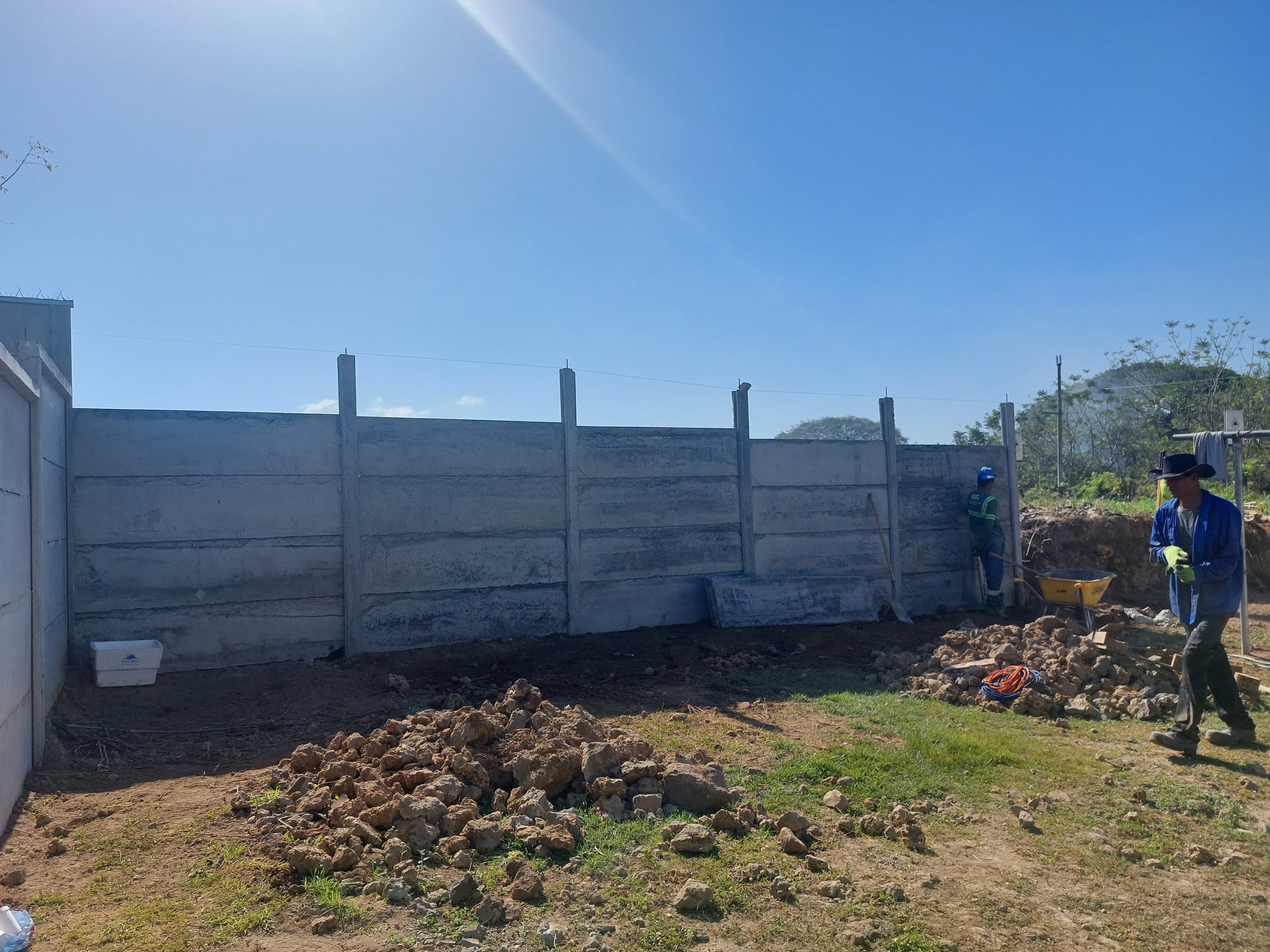 Muro perimetral prefabricado de concreto en lote rural con cuadrilla preparando el terreno al fondo