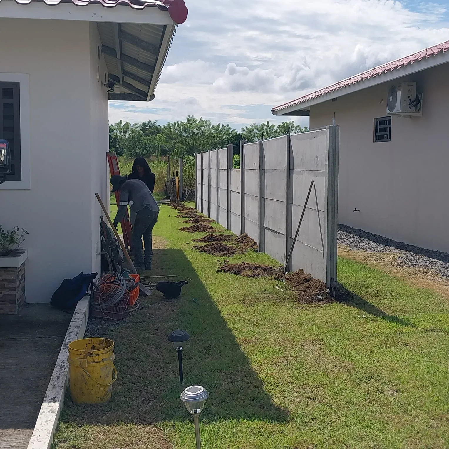 Cuadrilla instalando muro perimetral lateral junto a vivienda con techo de teja y poste solar
